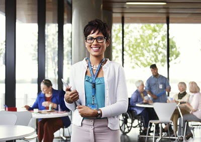 young female professional (student) holding clipboard, with seniors and a health care professional in the background.